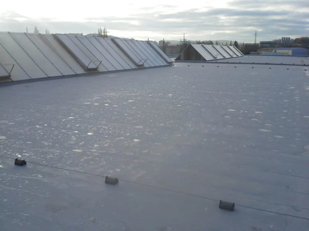 Plochá střecha budovy pokrytá šedou hydroizolační fólií, s proskleným světlíkem v rohu, obklopená otevřenou krajinou a poli.