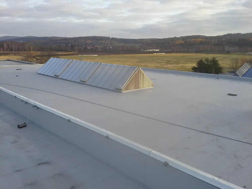 Plochá střecha průmyslové budovy s bílým světlíkem a technickým zařízením, v pozadí podzimní krajina s domy a horským hřebenem.