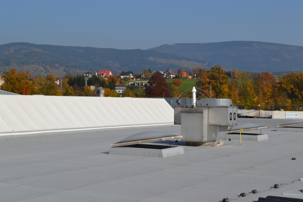 Plochá střecha průmyslové haly s několika světlíky a technickou jednotkou v pozadí panelová sídliště a stromy.
